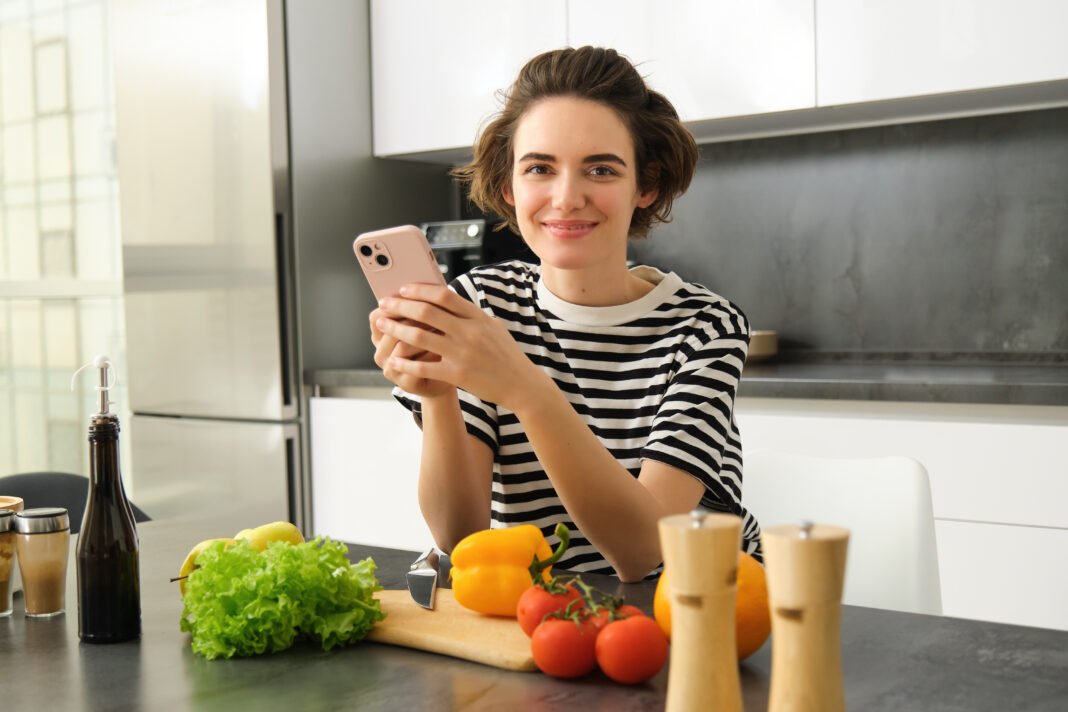 Portrait of young woman standing in the kitchen with vegetables and chopping board, holding smartphone, searching healthy diet recipes on mobile phone browser, cooking at home 5 influenciadores de culinária